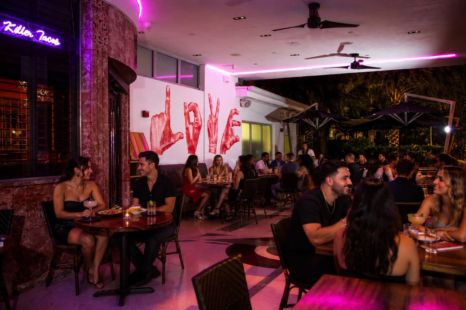 Guests with margaritas on the covered outdoor patio at Naked Taco — neon signage, the LOVE mural and umbrella seating on Collins Avenue, South Beach